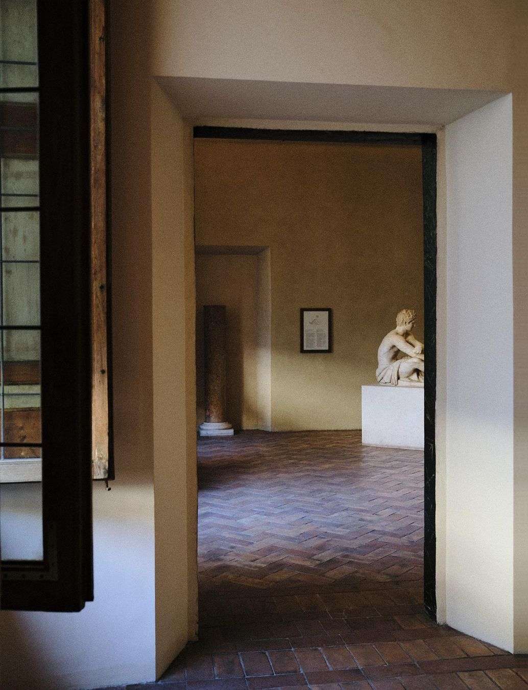 Interior view through doorway of art gallery with sculpture of seated figure on plinth, brick floor, windows and natural light.