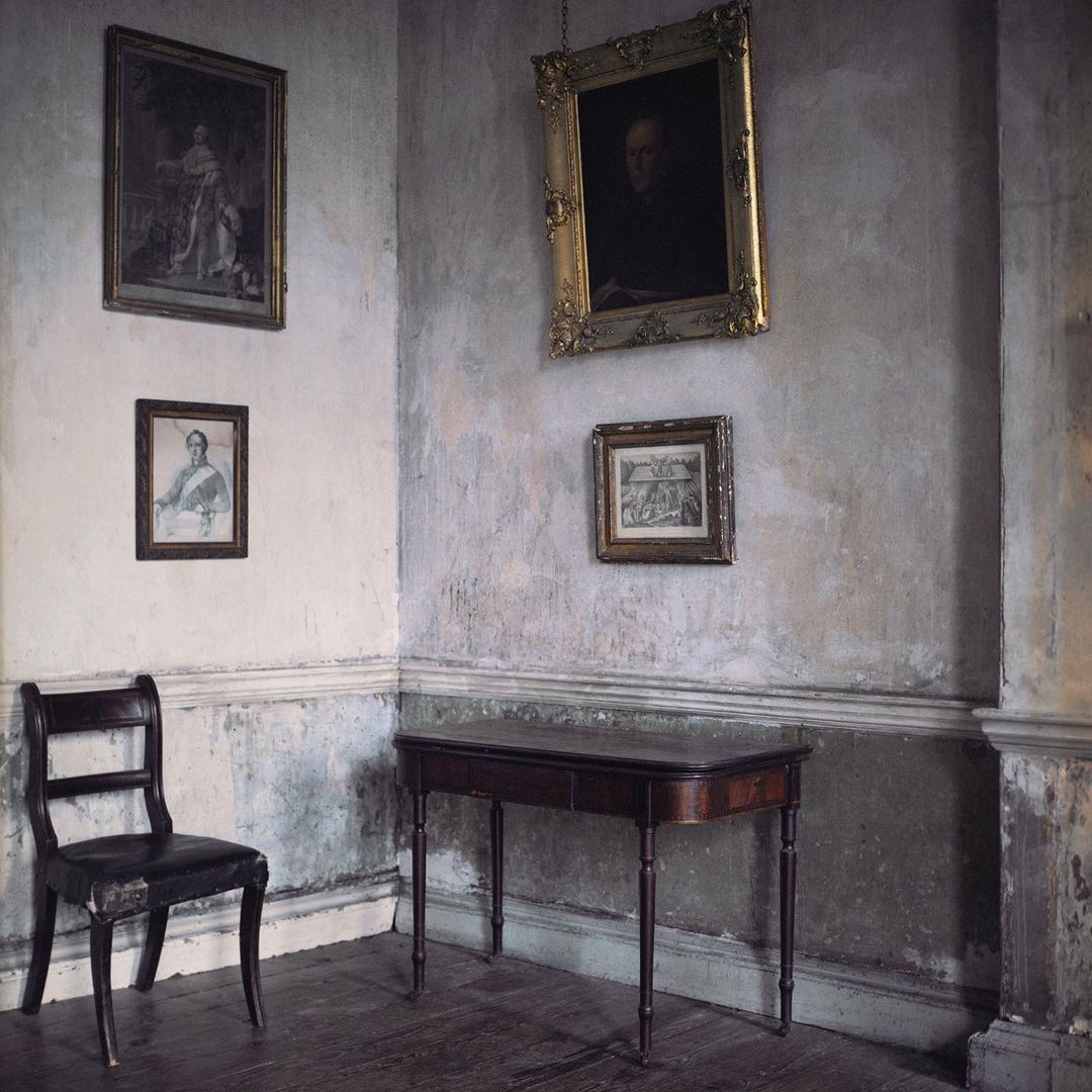 Interior of a room featuring a wooden chair and table, with vintage framed portraits hanging on the textured, grey-toned walls.