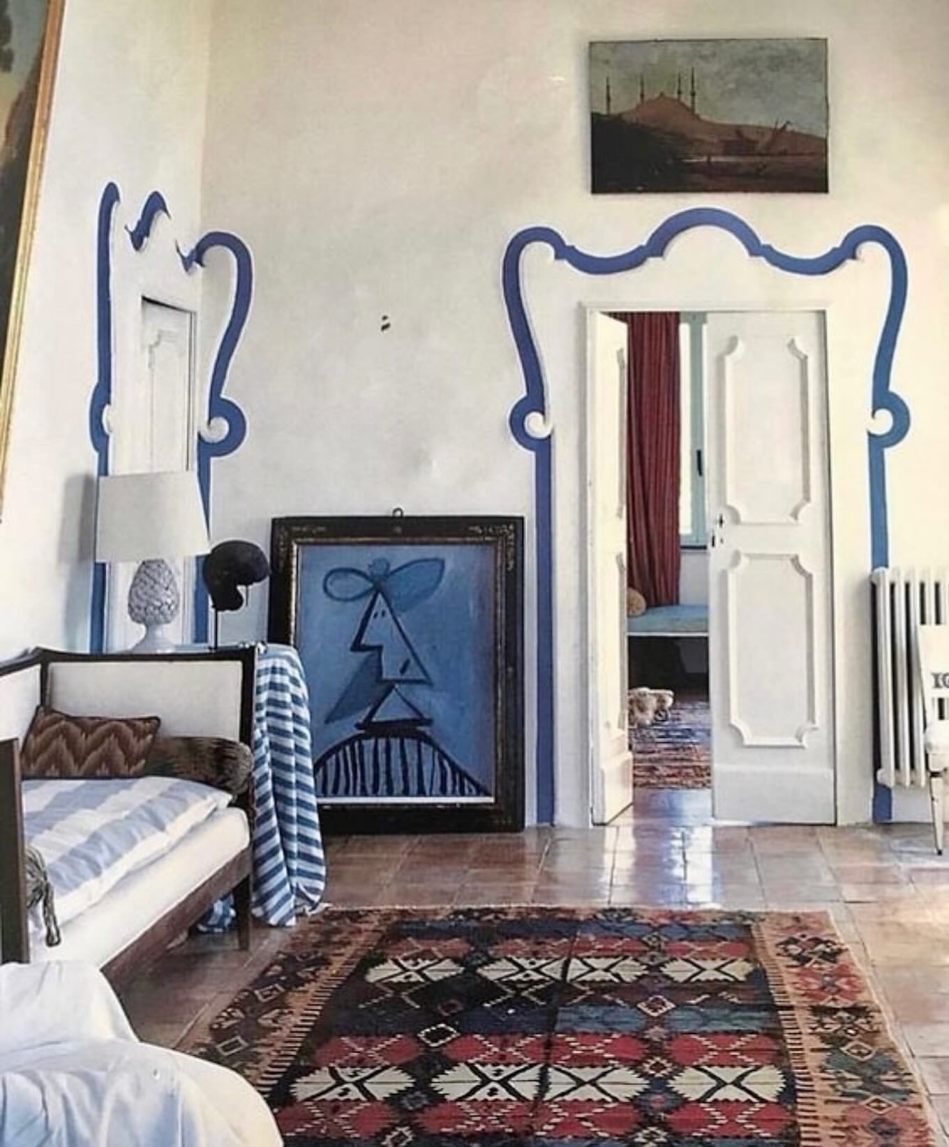 Interior of a room featuring a bed, patterned rug, an abstract painting of a face, and architectural details like a doorway.