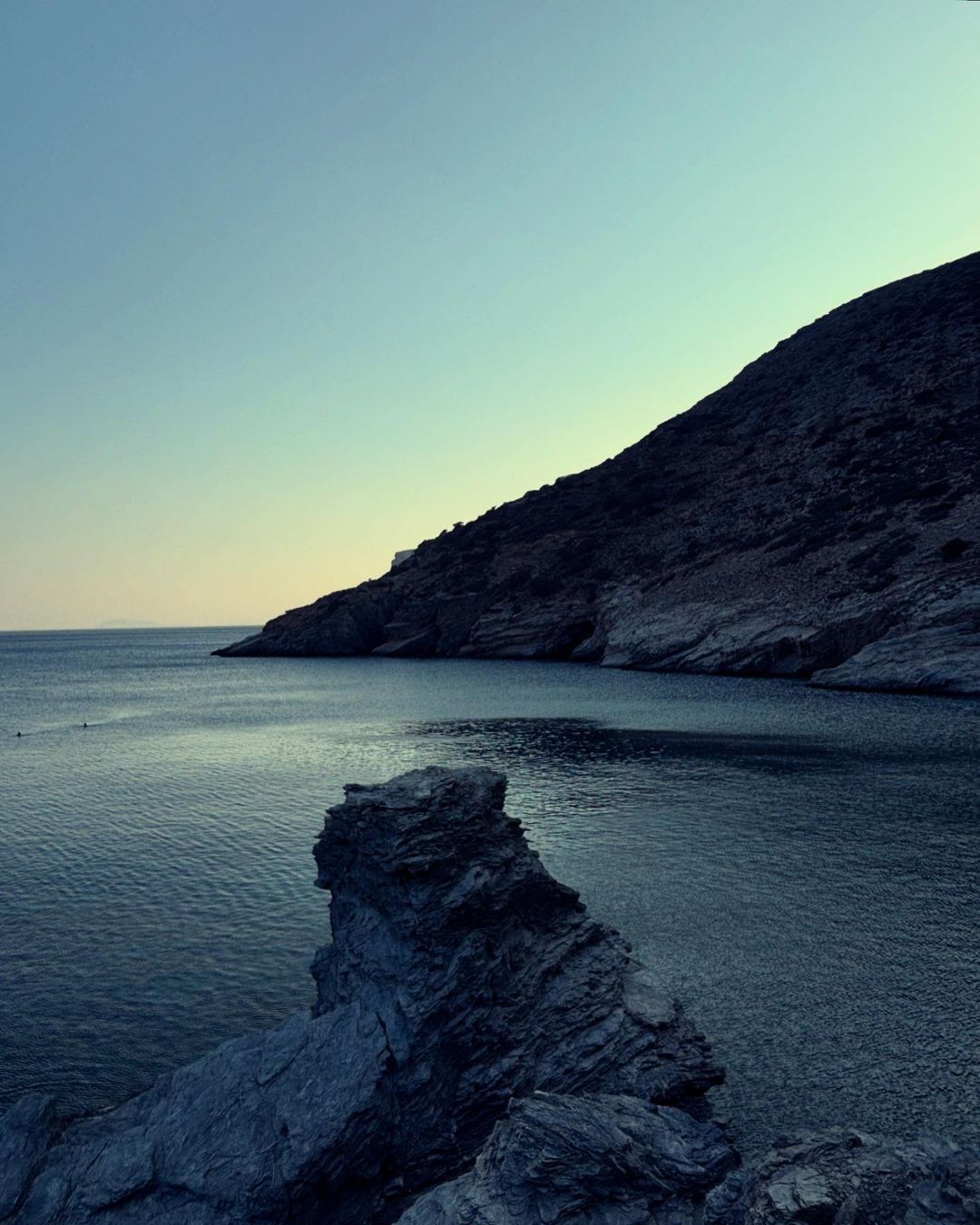 Seascape view featuring dark blue water, rugged rocks in the foreground, and a hill in the background under a blue sky.