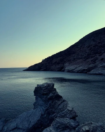 Seascape view featuring dark blue water, rugged rocks in the foreground, and a hill in the background under a blue sky.
