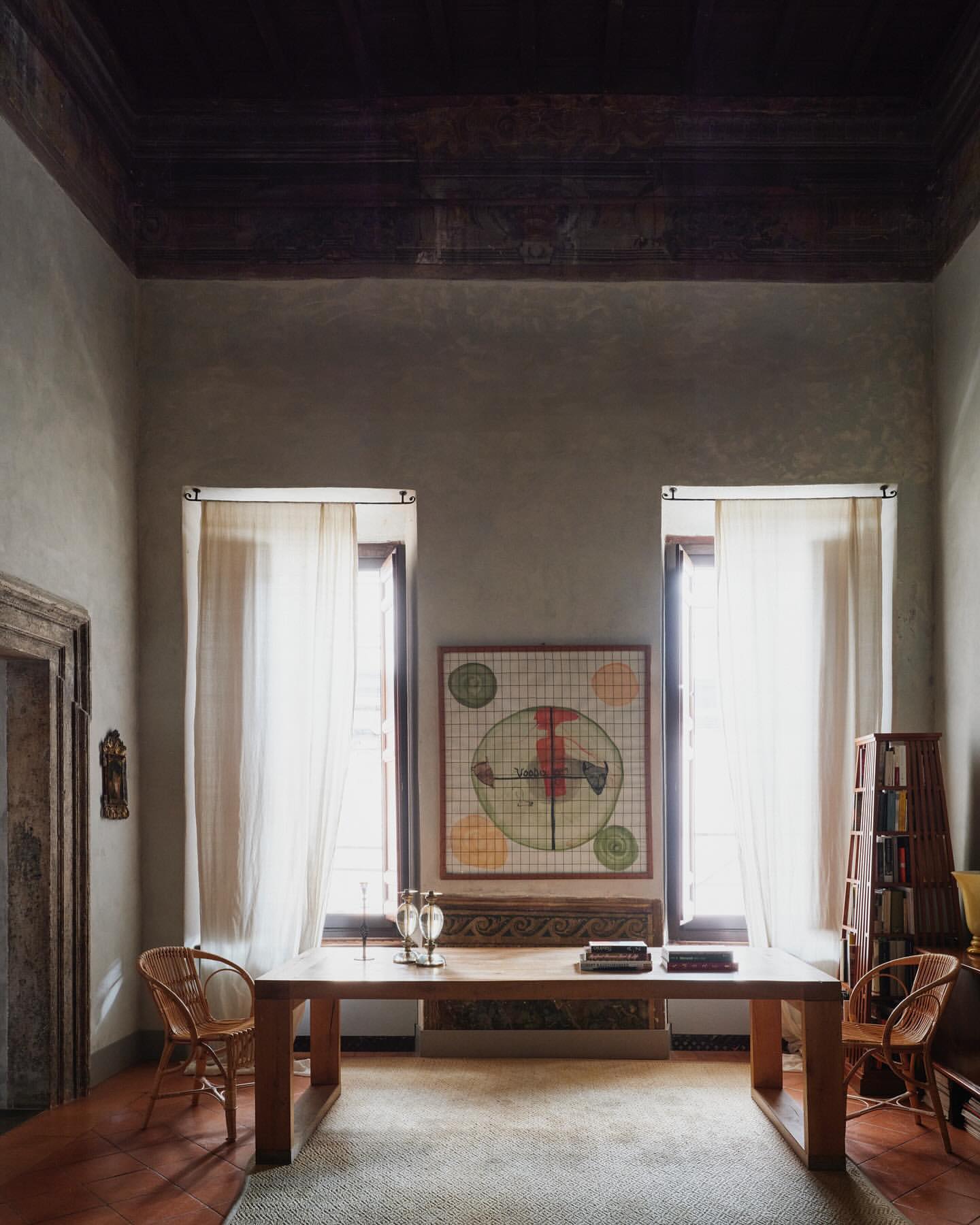 Interior view of a living room with a wooden table, artwork, and windows with sheer curtains, creating a bright, inviting space.
