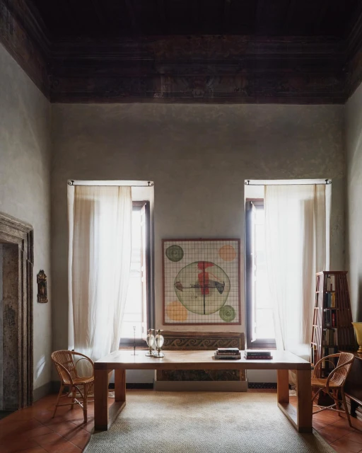 Interior view of a living room with a wooden table, artwork, and windows with sheer curtains, creating a bright, inviting space.