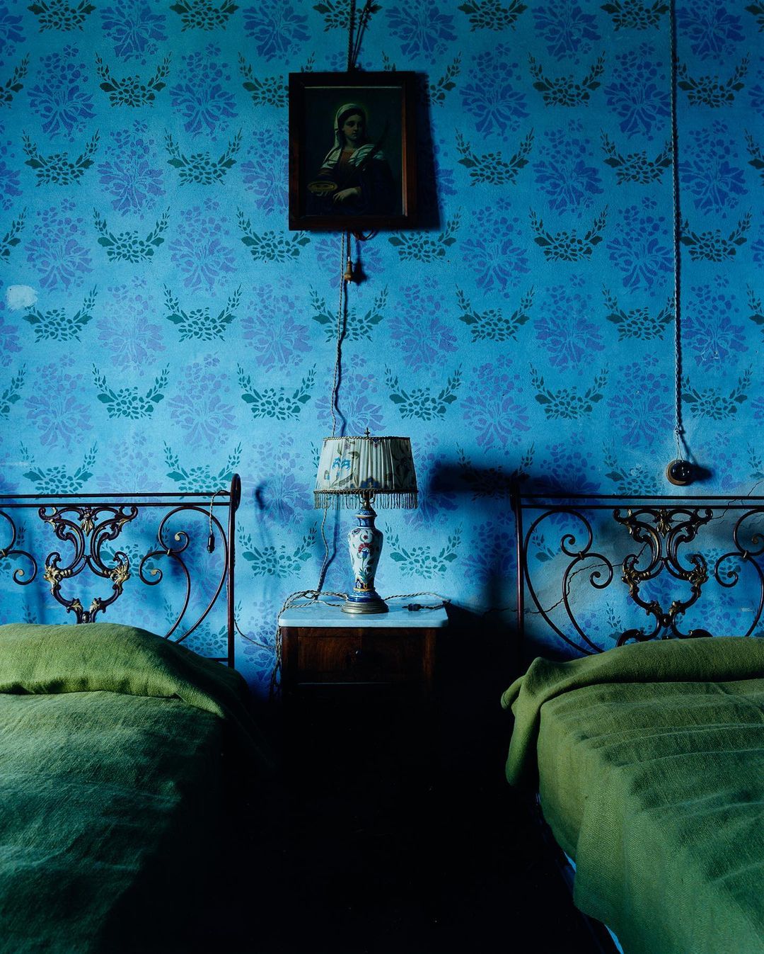 Vintage bedroom with twin beds featuring olive green blankets, ornate metal headboards, and floral patterned wallpaper in shades of blue.