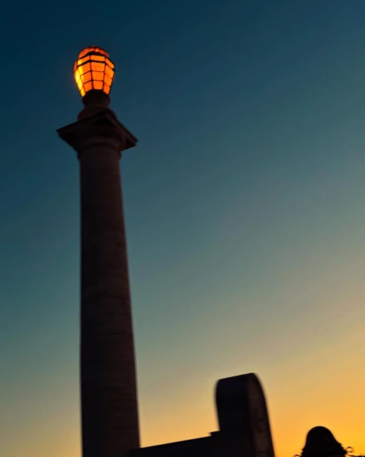 Illuminated monument with a light fixture atop a tall column, set against a gradient sky from orange to blue at dusk or dawn.