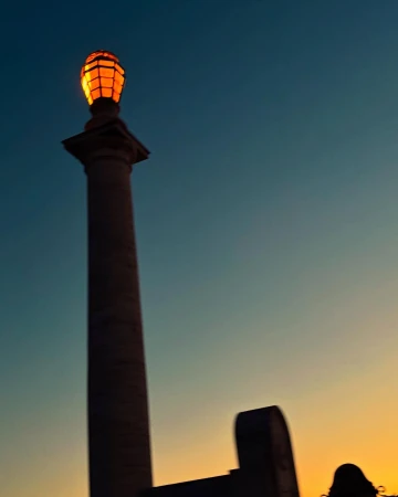 Illuminated monument with a light fixture atop a tall column, set against a gradient sky from orange to blue at dusk or dawn.