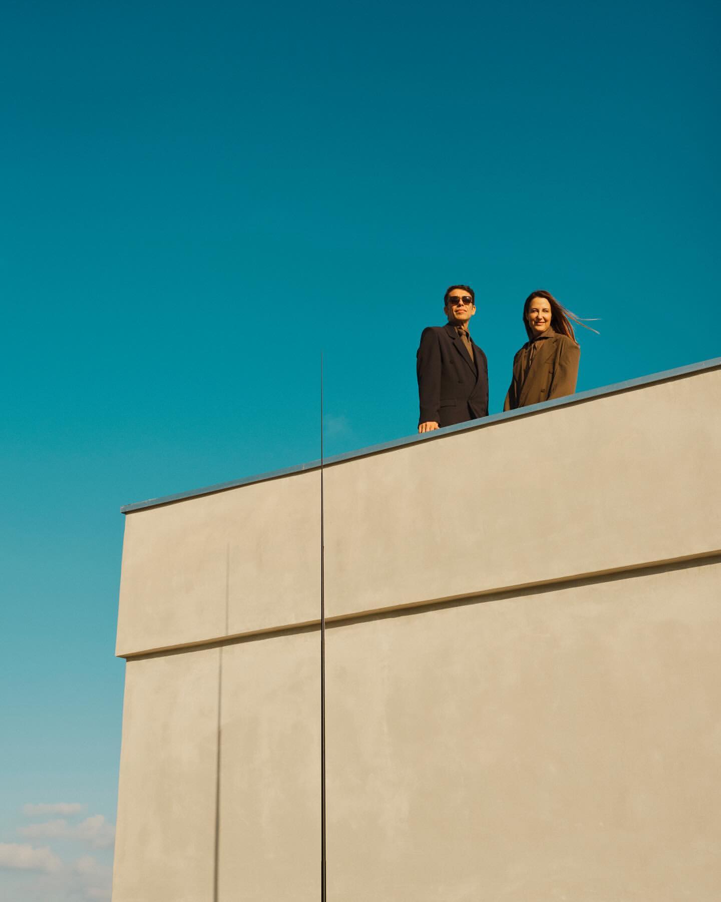 Two people standing on a rooftop ledge against a bright blue sky, photographed by Simon Watson.