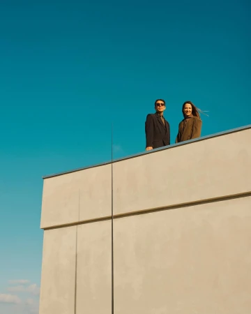 Two people standing on a rooftop ledge against a bright blue sky, photographed by Simon Watson.