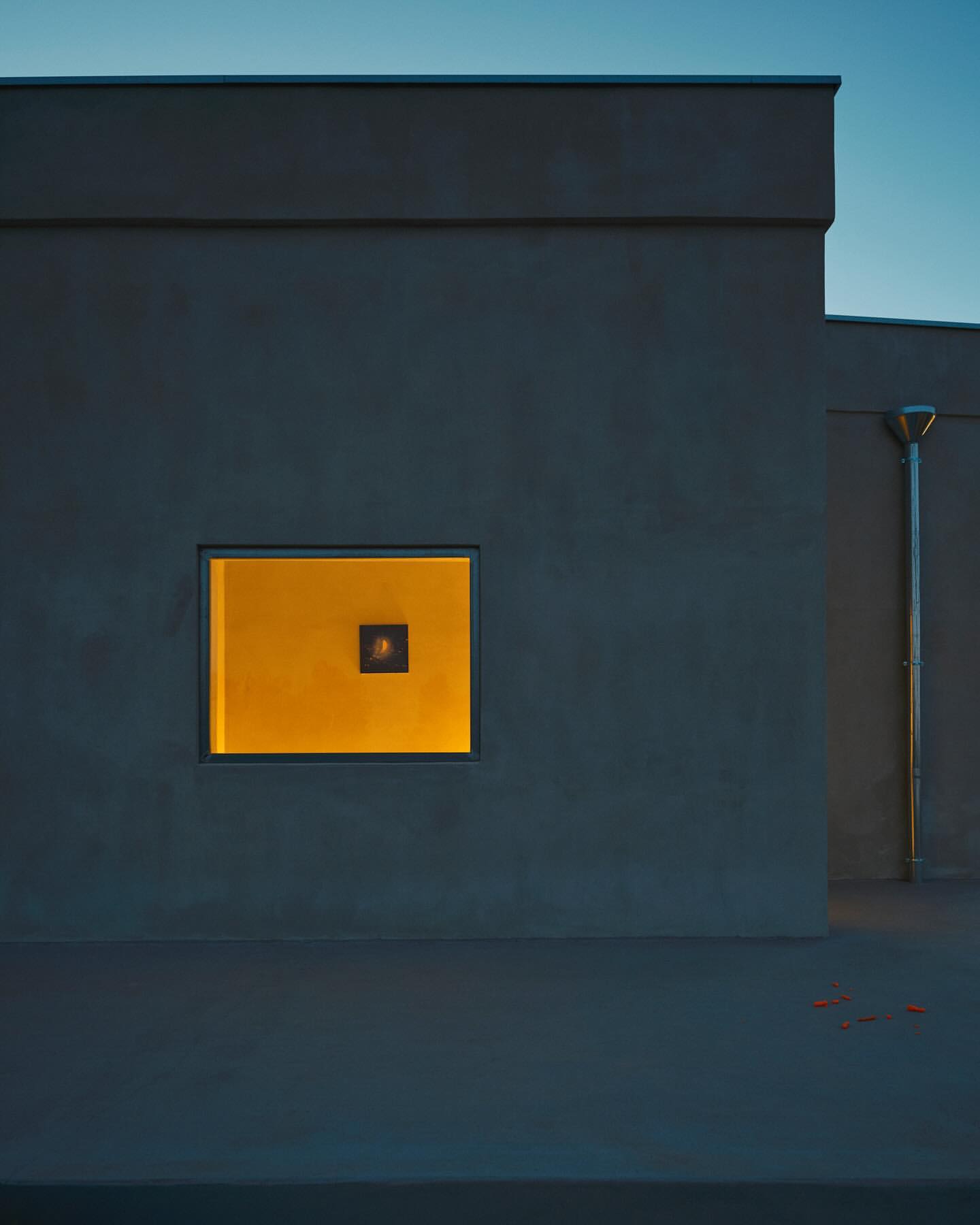 Architectural facade at dusk with a square window emitting golden light, set against a muted blue sky and dark building exterior.
