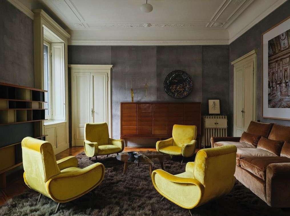Living room interior with four yellow armchairs on a brown rug, a brown sofa, wooden cabinet, and white walls with white trim.