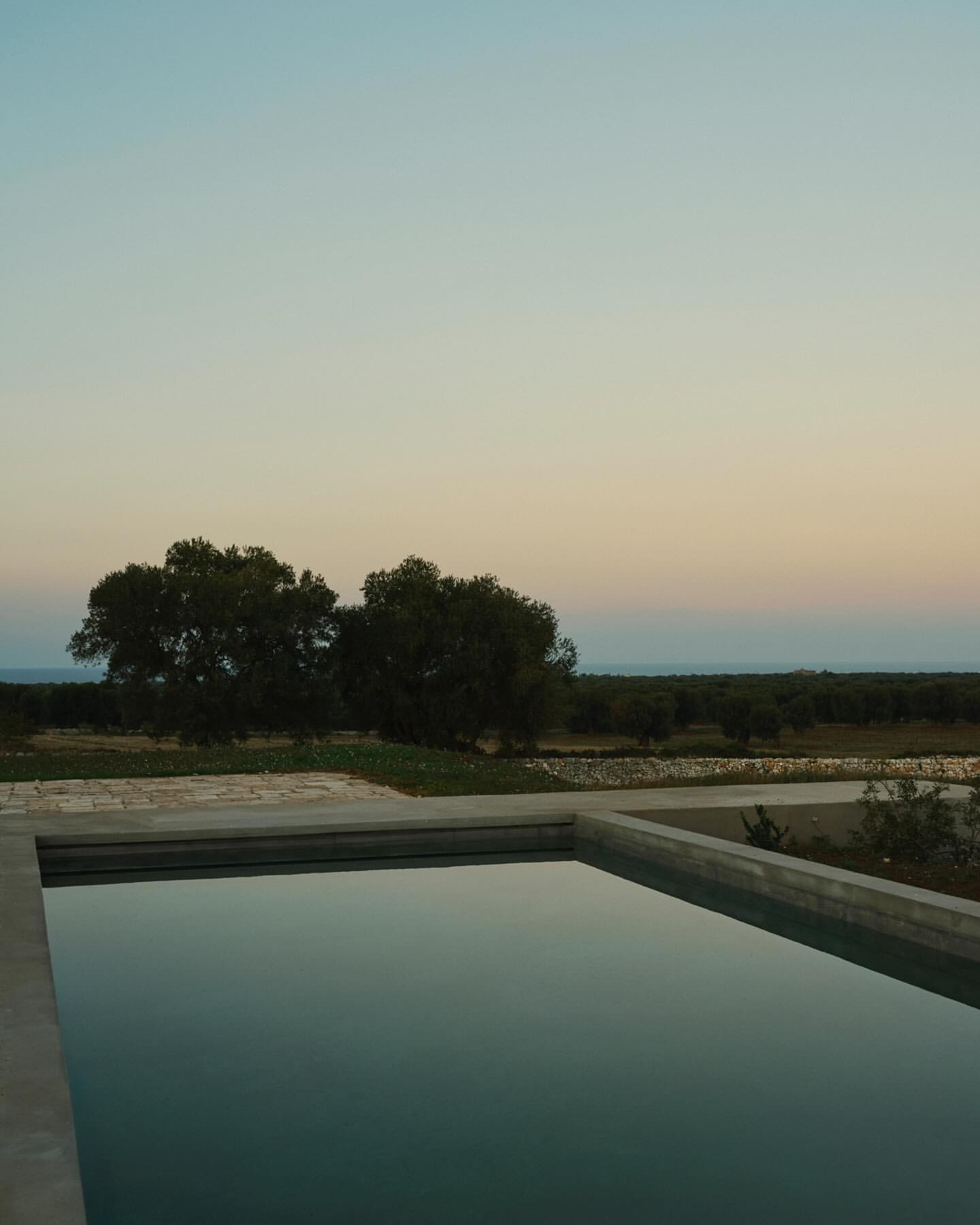 Still shot of a concrete swimming pool with calm water reflecting the evening sky, trees and distant landscape beyond the pool edge.