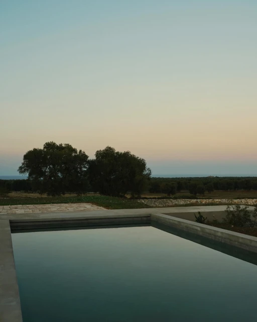 Still shot of a concrete swimming pool with calm water reflecting the evening sky, trees and distant landscape beyond the pool edge.