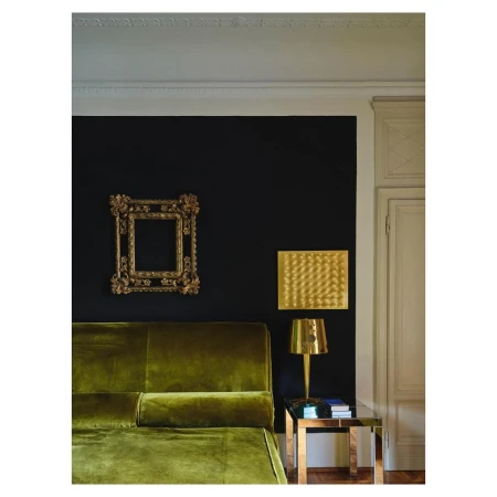 Interior with olive-green velvet couch, gold ornate mirror frame on black wall, and gold lamp on glass and gold side table.