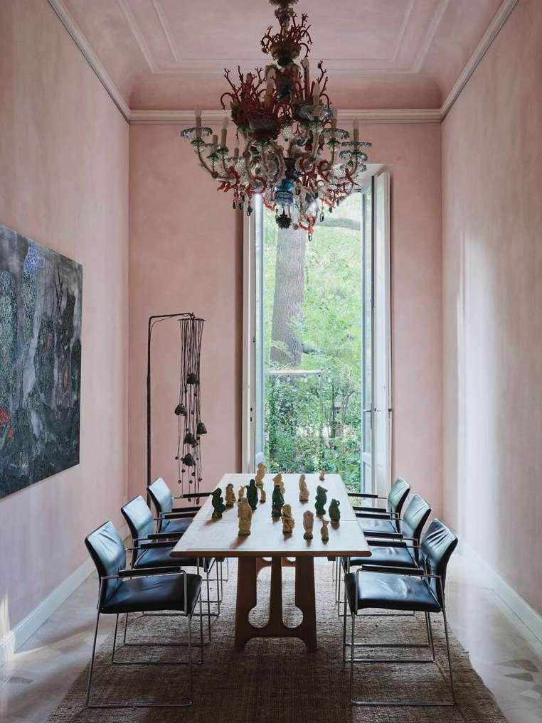 Dining room interior with a table set with figurines, surrounded by chairs and lit by an ornate chandelier, in a room with pink walls.
