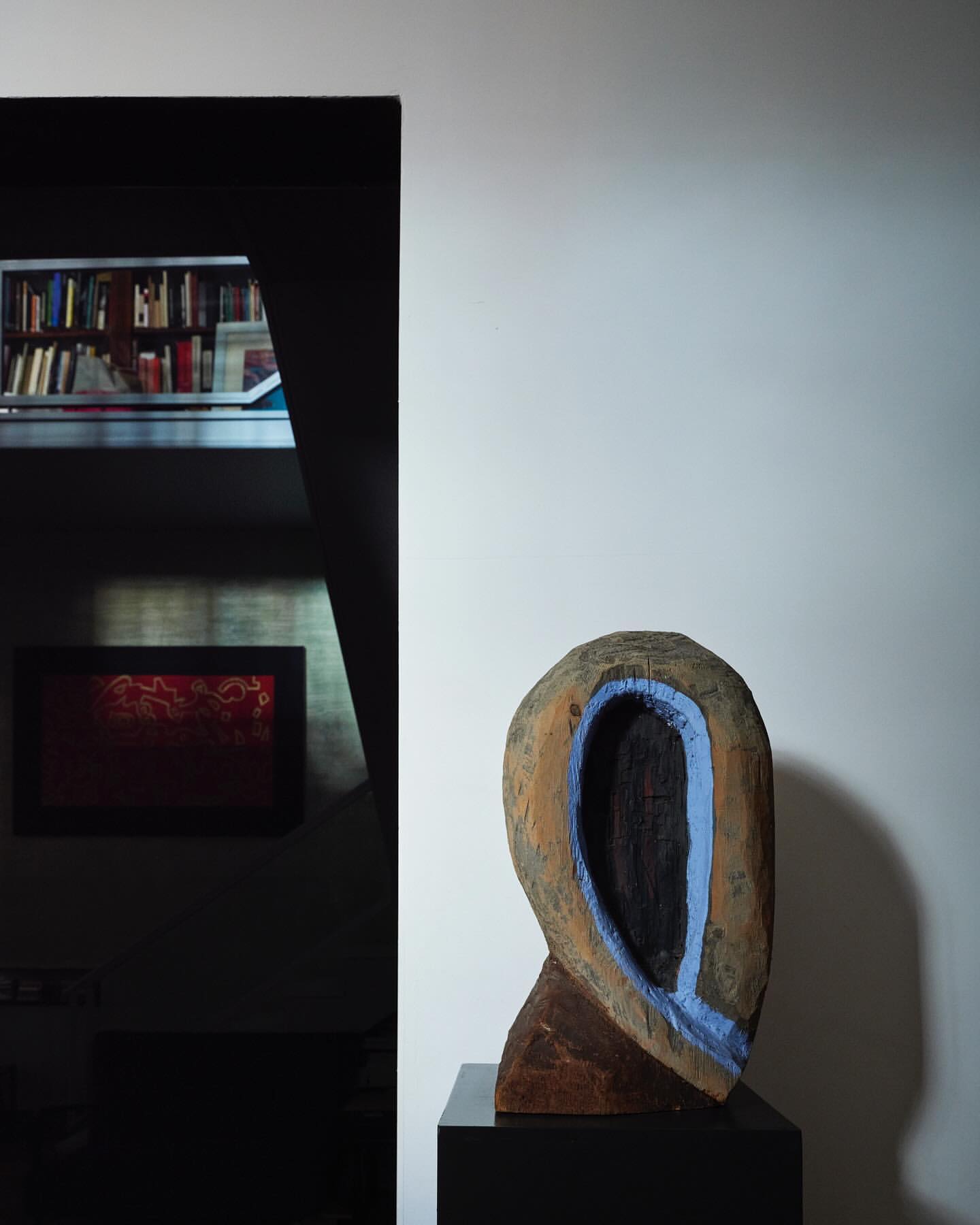 A wooden art sculpture stands on a black pedestal in a room with a bookshelf in the background, creating a minimalist art display.