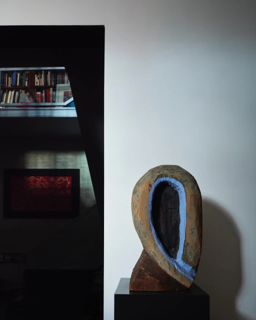A wooden art sculpture stands on a black pedestal in a room with a bookshelf in the background, creating a minimalist art display.