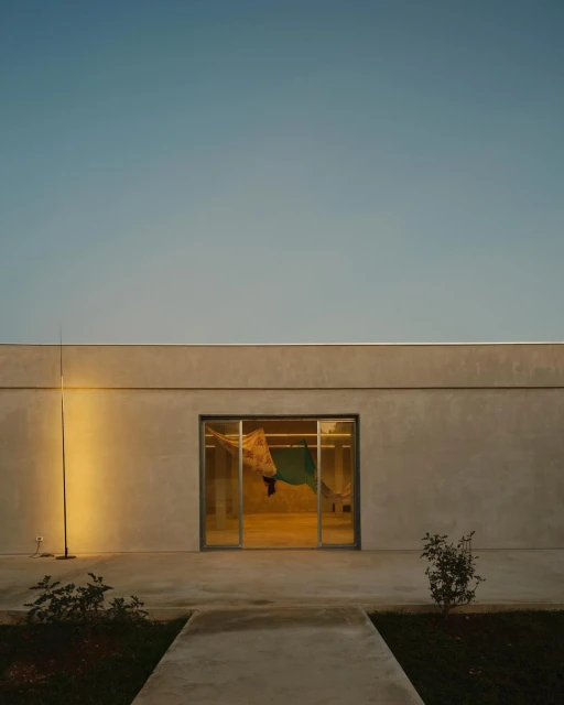 Modern architectural building with a minimalistic design, featuring a neutral color palette and a large window, surrounded by manicured greenery.