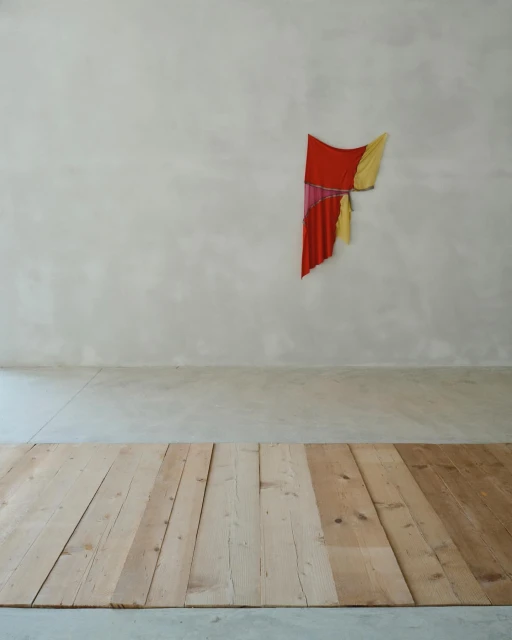Minimalist art installation featuring draped red, yellow, and pink fabrics on a gray wall, complemented by a light-toned wooden plank floor.