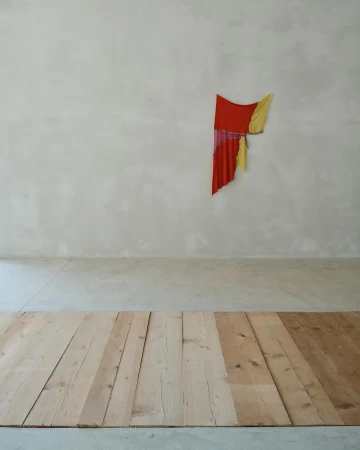 Minimalist art installation featuring draped red, yellow, and pink fabrics on a gray wall, complemented by a light-toned wooden plank floor.
