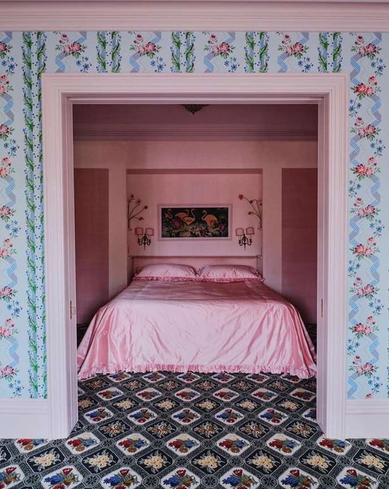 A bedroom viewed through a doorway with pink molding, featuring a pink bed and floral patterned wallpaper.