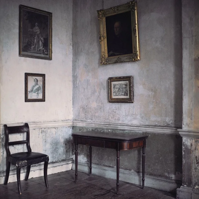 A rustic room corner with distressed plaster walls, antique framed portraits, a dark wooden chair, and a slim side table.