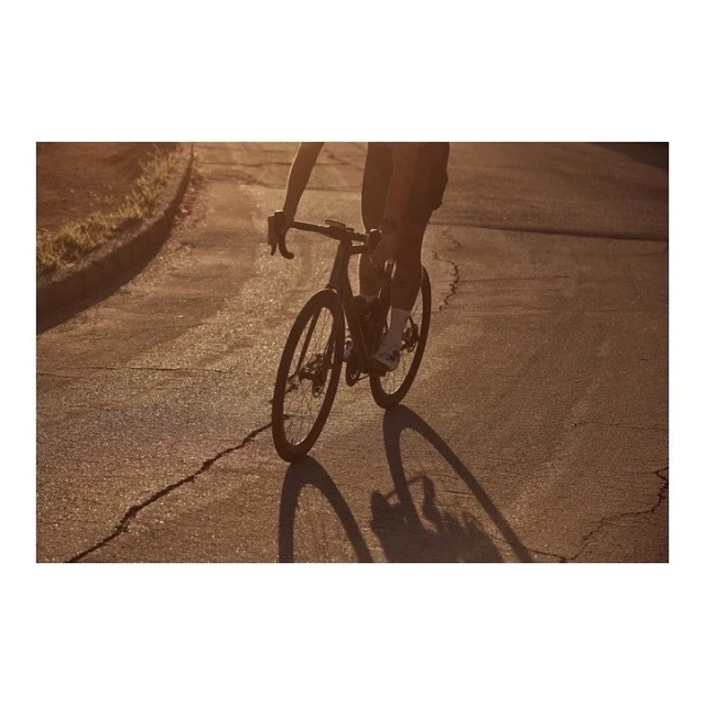 An adult rides a road bike on a cracked asphalt road in the late afternoon or early morning light, casting a long shadow.