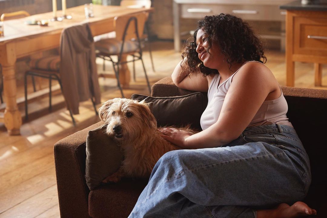 Woman smiles on a brown couch with a terrier mix dog. Light streams into the home from a window behind the wooden dining table.
