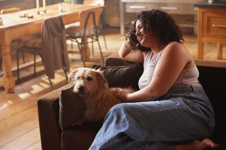 Woman smiles on a brown couch with a terrier mix dog. Light streams into the home from a window behind the wooden dining table.