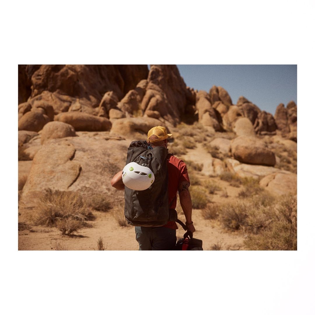 Man hikes in desert landscape with backpack, climbing helmet, and hat, walking away from the camera with rocky hills in background.