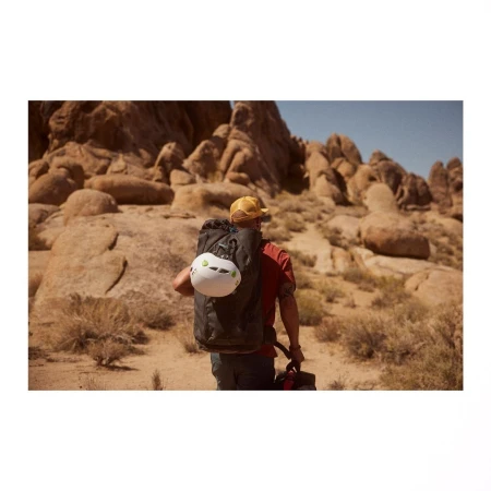 Man hikes in desert landscape with backpack, climbing helmet, and hat, walking away from the camera with rocky hills in background.