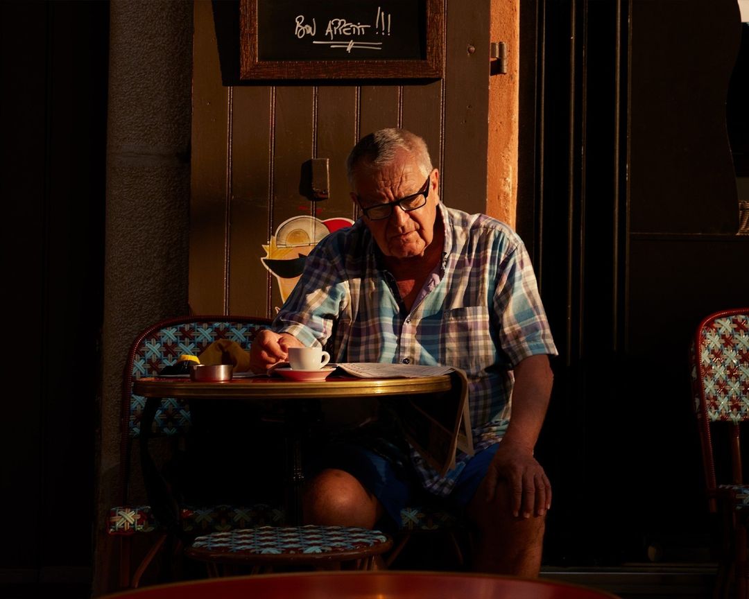 Elderly man sits alone at outdoor cafe table, reading newspaper and drinking coffee in warm light. Quaint scene with European charm.