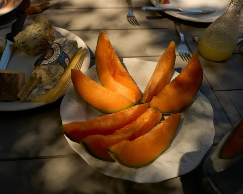 Overhead shot of a plate of melon slices, cheese, and a beverage on a wooden table, with natural sunlight creating dynamic shadows.