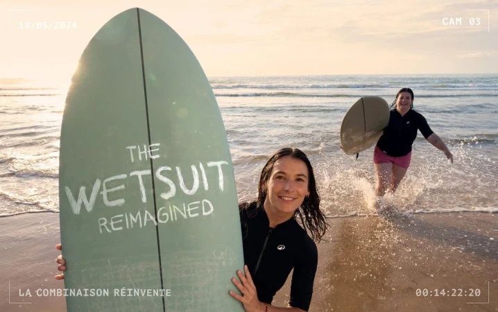 Two women holding surfboards on a sandy beach at sunset, captured in a wide horizontal coastal scene with gentle waves.