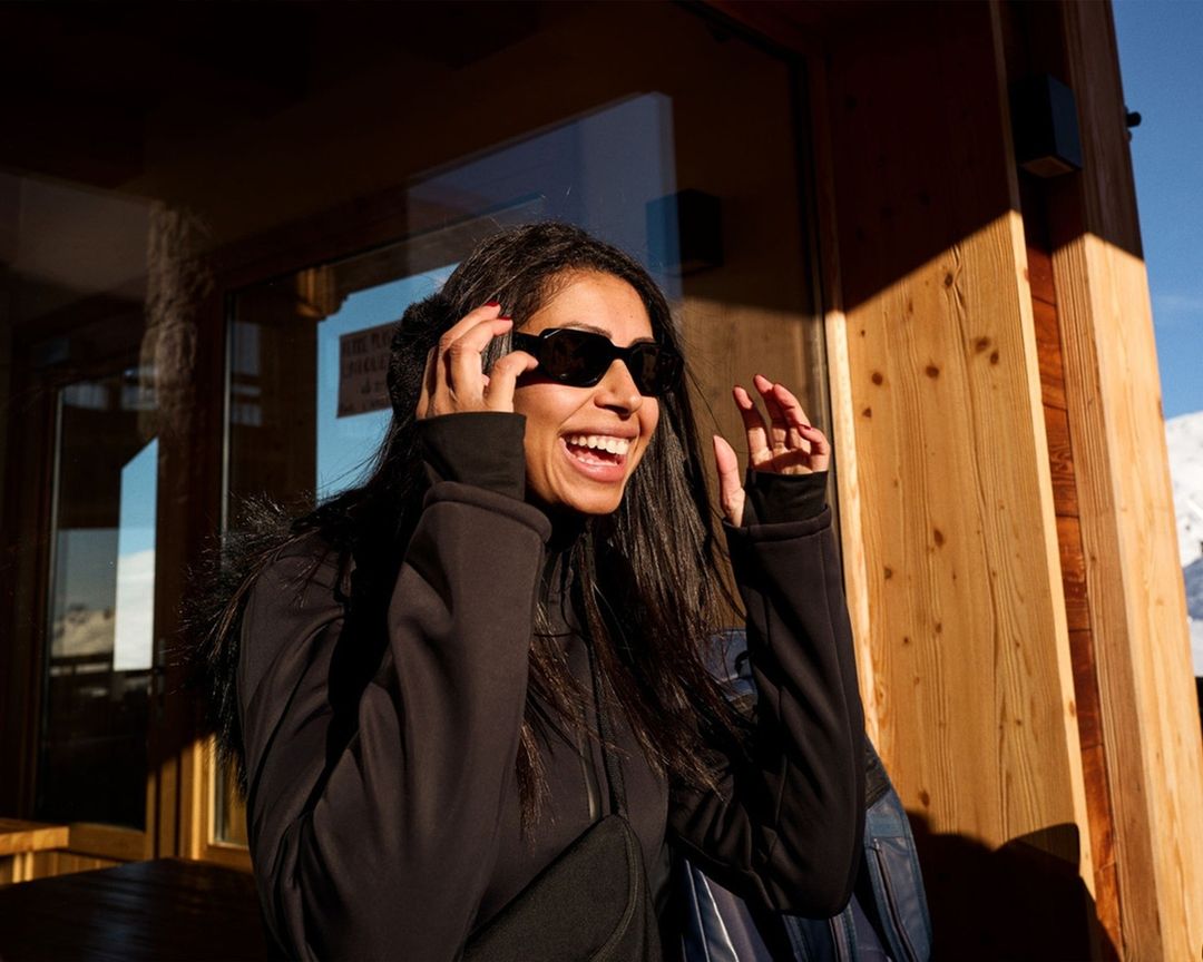 Smiling woman with sunglasses standing in front of a wooden building on a sunny day in a snowy, mountainous area with blue sky.