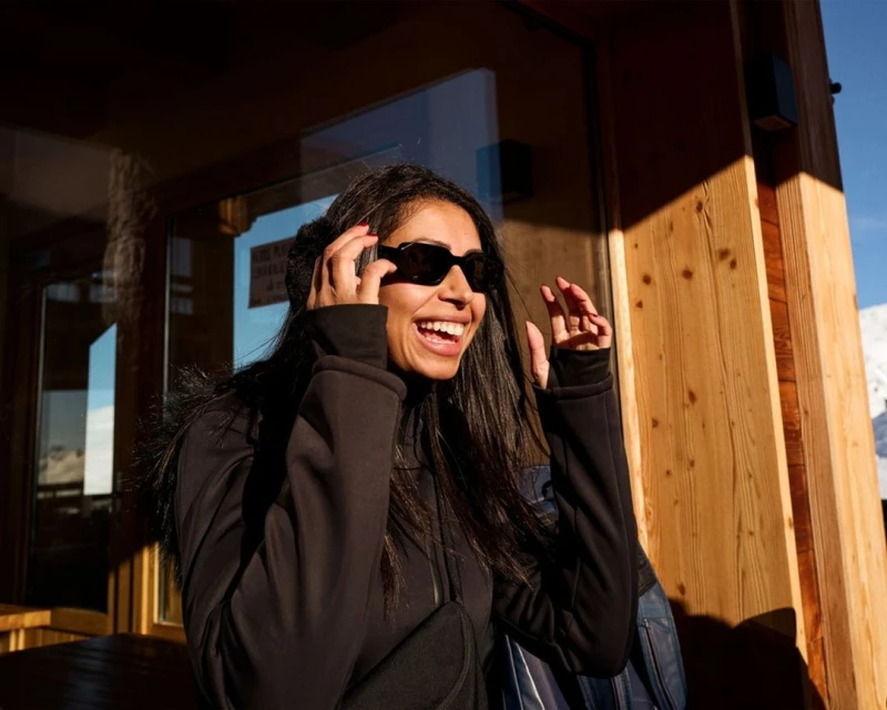 Smiling woman with sunglasses standing in front of a wooden building on a sunny day in a snowy, mountainous area with blue sky.