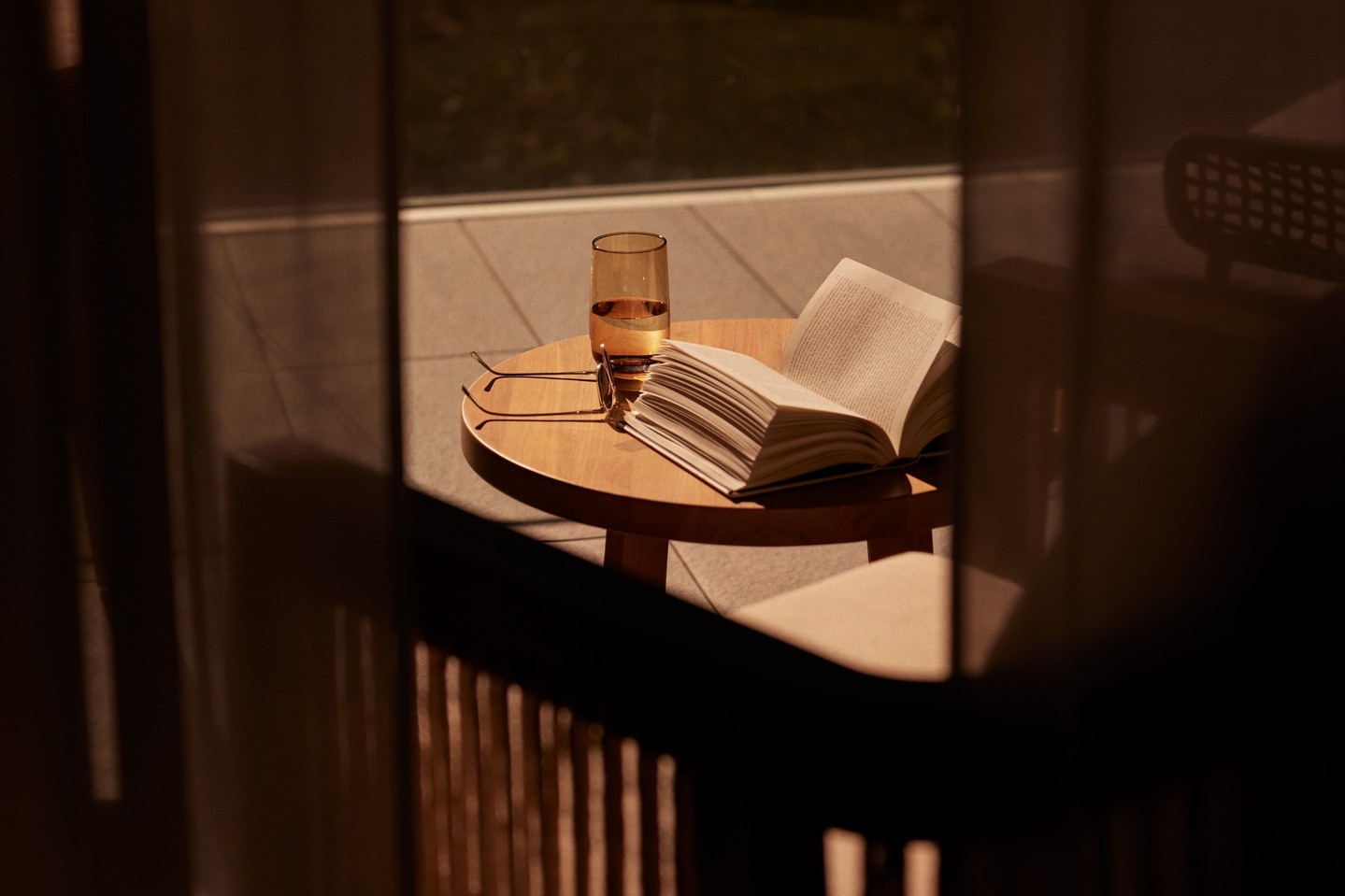Still life with open book, glasses, and drink on a round table, seen through a window with filtered light and outdoor background.