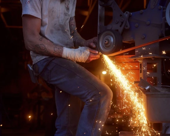 Adult male at work grinds metal with a belt grinder, producing sparks in a dimly lit workshop, wearing safety bandages and blue jeans.