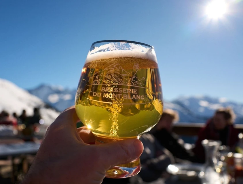 Close-up of a hand holding a glass of beer in front of a mountain range, with a sunny sky, showcasing a beer product in a natural setting.