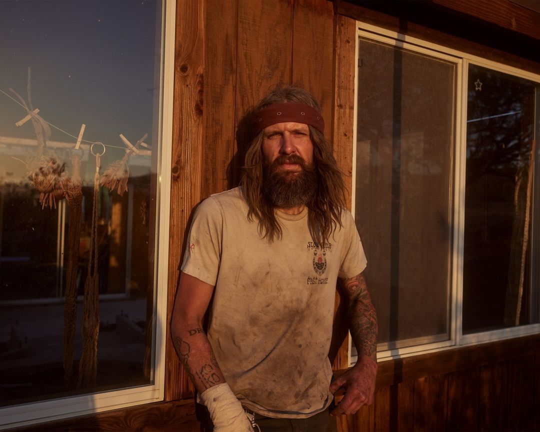 Portrait of a bearded man with long hair and a bandana standing in front of a wooden building with a window reflecting the sunset.