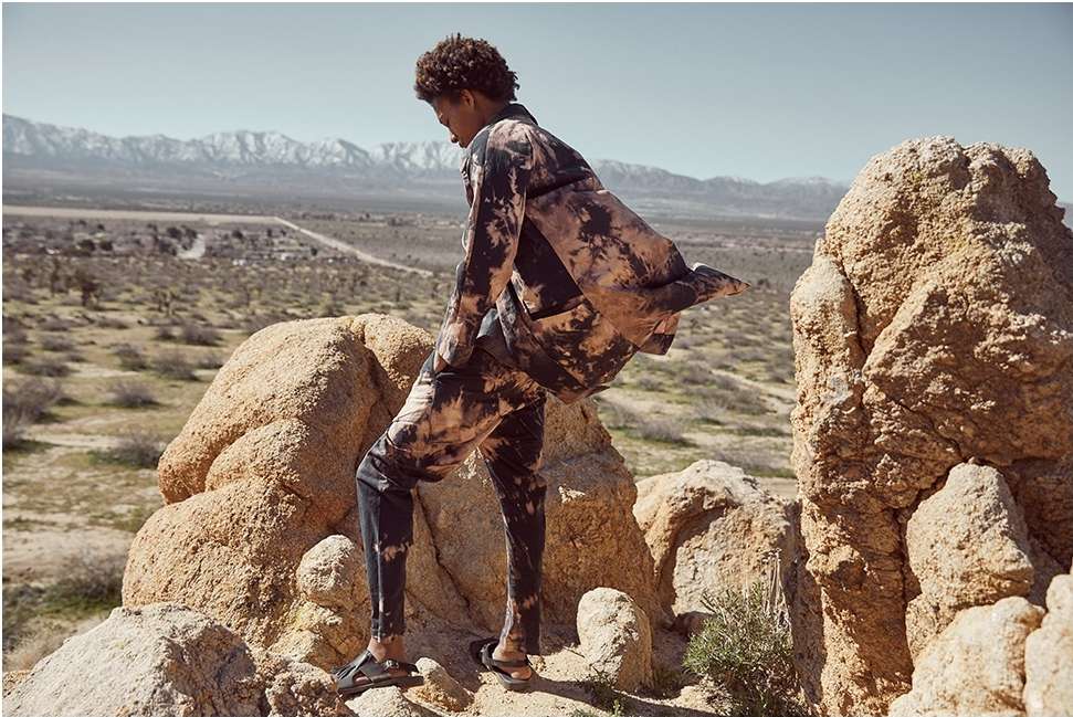 Male model in tie-dye suit poses on desert rocks. Arid landscape, mountains, and sky in the background create a scenic backdrop.