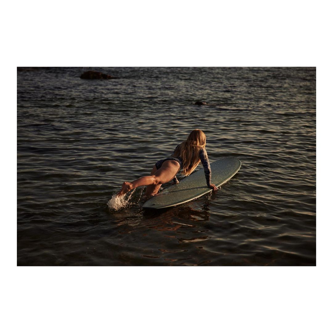 Woman in a swimsuit lying on a surfboard in the water, with her feet splashing in the sea, ocean background, and evening light.