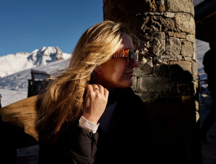 Woman in sunglasses poses near stone wall with snow-covered mountains in the background on a sunny day, captured in profile.