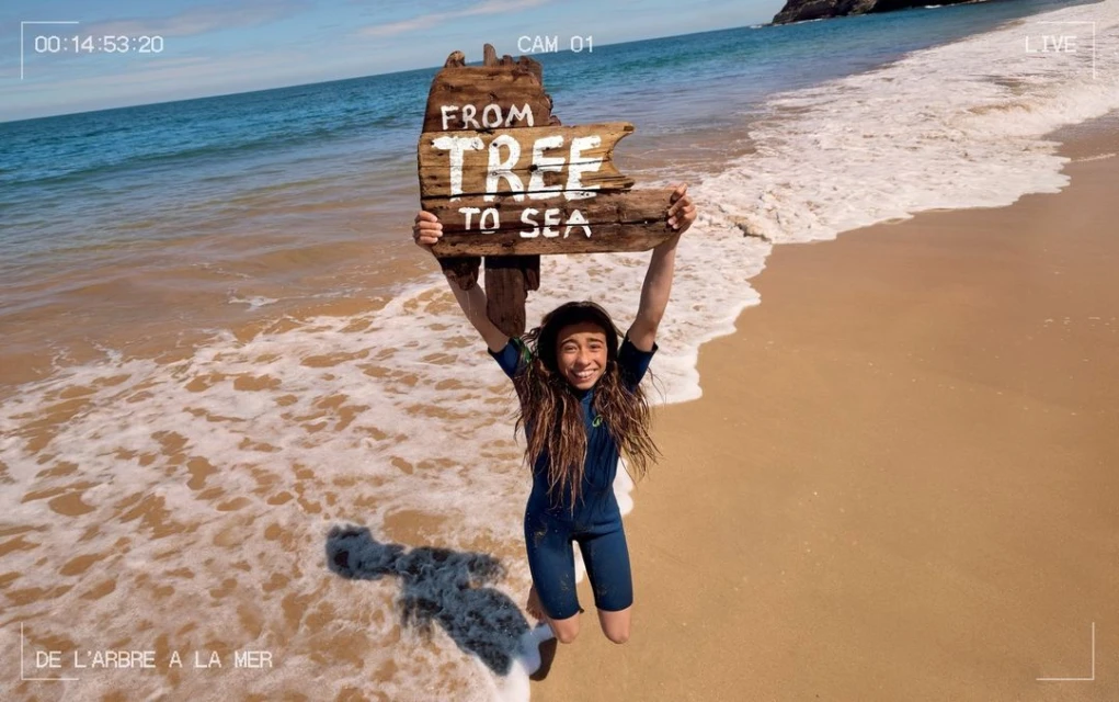 A young person in a wetsuit gleefully holds a wooden sign saying 'From Tree to Sea' above their head on a sandy beach by the ocean.