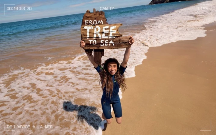 A young person in a wetsuit gleefully holds a wooden sign saying 'From Tree to Sea' above their head on a sandy beach by the ocean.