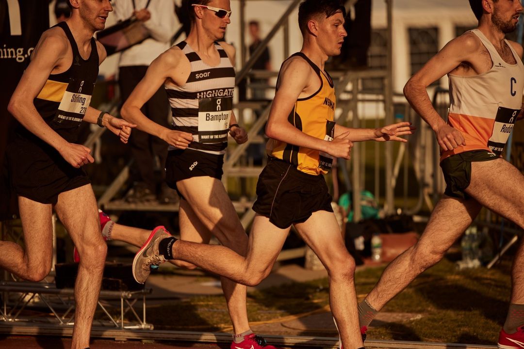 Four athletes compete in an outdoor track and field race on a sunny day. Runners wear black shorts and racing tops with sponsor logos.