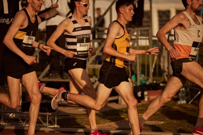 Four athletes compete in an outdoor track and field race on a sunny day. Runners wear black shorts and racing tops with sponsor logos.