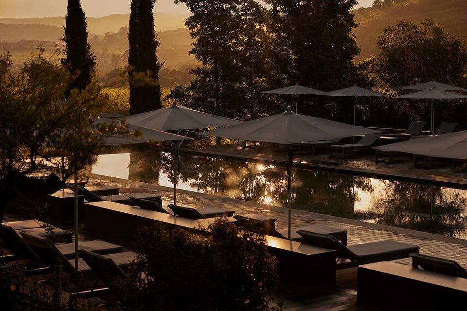 Outdoor resort pool with lounge chairs and umbrellas reflects the golden light of the setting sun in a scenic, hillside setting.