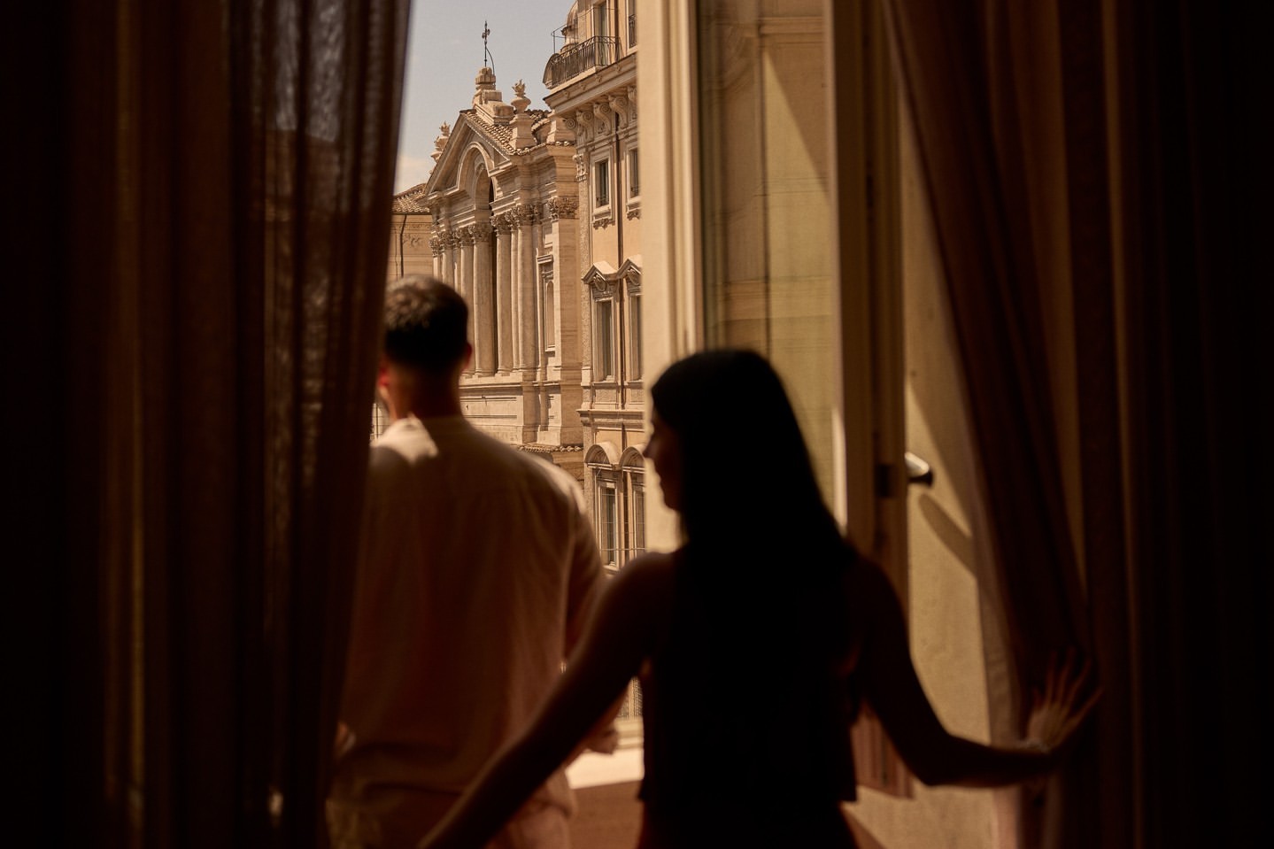 Back view of a man and a woman standing at a window overlooking architecture with columns and a dome, framed by open curtains.