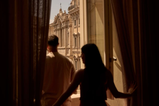 Back view of a man and a woman standing at a window overlooking architecture with columns and a dome, framed by open curtains.