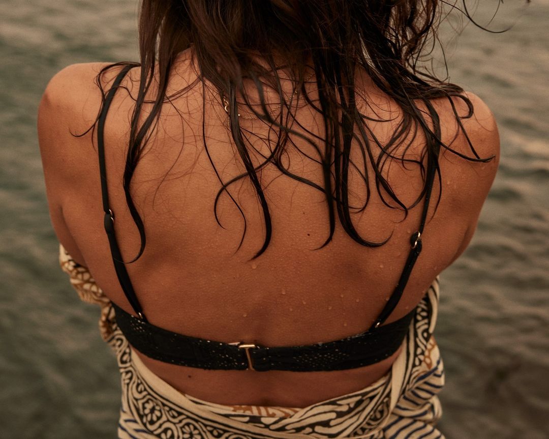 Woman with wet dark hair, black bikini top, and patterned sarong seen from the back against a water background on a cloudy day.